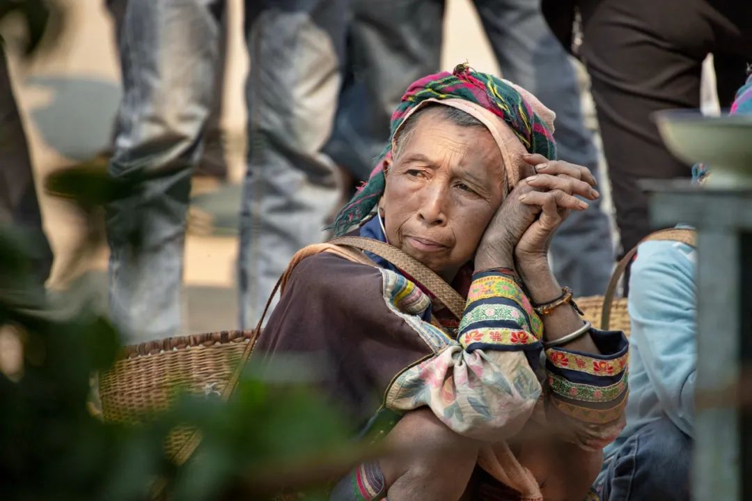 蹲坐的一位拉祜族老妇人,她的身后是几个穿着牛仔裤的少年.